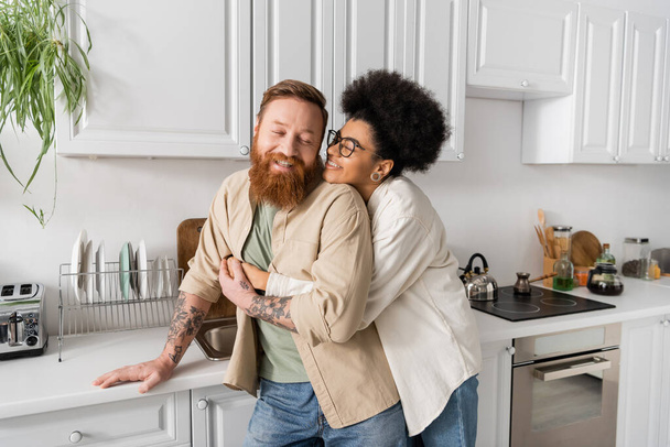 Cheerful african american woman hugging tattooed boyfriend in kitchen  - Foto, Imagem