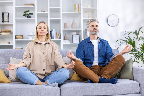 Dorosła para mężczyzna i kobieta relaksujący się razem w domu na kanapie w pozycji lotosu, rodzina medytująca razem w salonie. - Zdjęcie, obraz