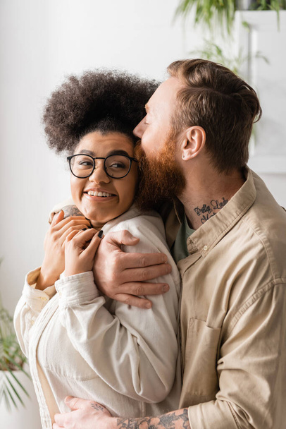 Bearded man kissing and hugging smiling african american girlfriend at home  - Foto, Imagen