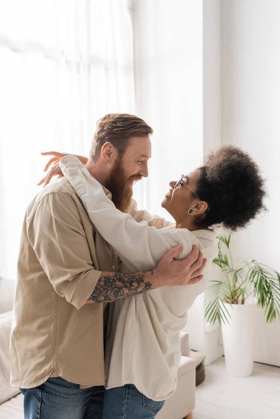 Side view of bearded man hugging and looking at happy african american girlfriend at home  - Foto, Imagem