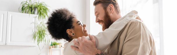 Side view of positive african american woman hugging bearded boyfriend at home, banner  - Photo, image