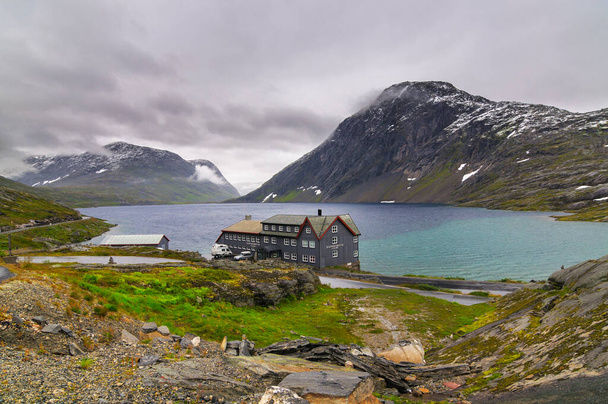 a hotel next to a lake in a fiord in norway with clouds in the sky and snow in the mountains - Photo, image