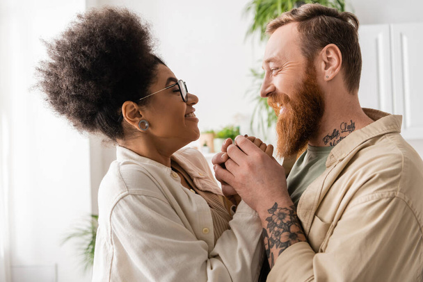 Side view of tattooed man holding hand of overjoyed african american girlfriend at home  - Фото, изображение