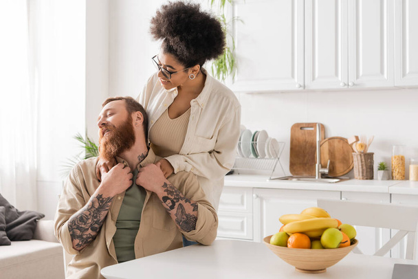 Positive african american woman hugging tattooed boyfriend in kitchen  - Фото, изображение