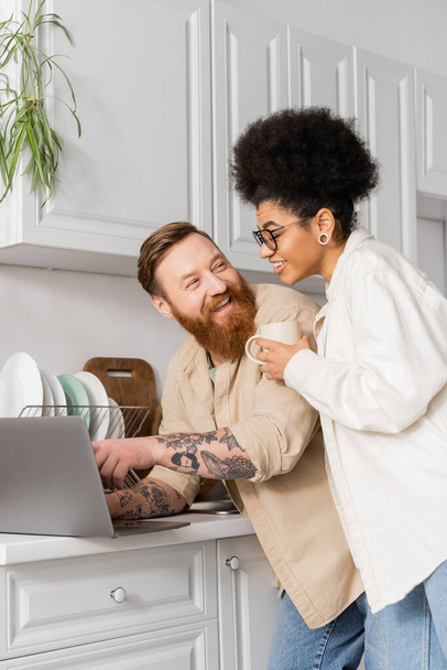 Tattooed man using laptop and looking at african american girlfriend with coffee in kitchen - Foto, Bild