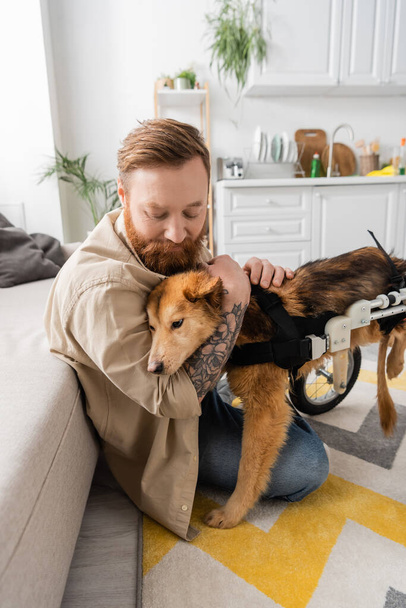Γενειοφόρος άντρας με τατουάζ αγκαλιάζει ανάπηρο σκύλο σε αναπηρική καρέκλα στο σπίτι  - Φωτογραφία, εικόνα