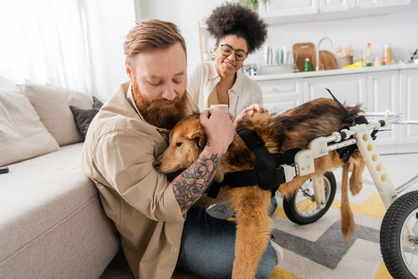 Tattooed man petting handicapped dog near african american girlfriend at home  - Foto, imagen