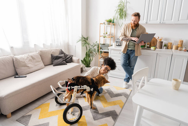 Smiling man using laptop near african american girlfriend and handicapped dog at home  - Foto, immagini