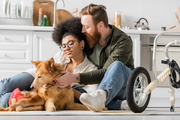 Tetovált férfi ölelés afro-amerikai barátnő és mozgássérült kutya közelében tolószék a konyhában  - Fotó, kép