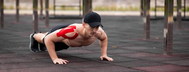 Young European athlete with injury is training outdoors. Kinesiology ( therapeutic, Kinesio, k-tape, KT, physio) tape on shoulder. Rehabilitation and recovery. Push up from the floor. - Fotó, kép