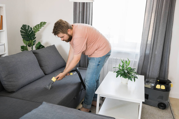 Mies, jolla on moderni pesu pölynimuri ja puhdistus likainen sohva ammattimaisesti pesuainetta. Professional springclean kotona - Valokuva, kuva
