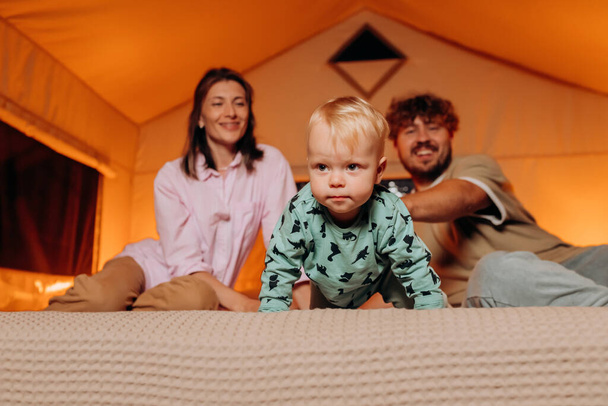 Happy family with lovely baby playing and spend time together in cozy glamping on summer evening while lying on bed. Luxury camping tent for outdoor recreation and recreation - Foto, Bild