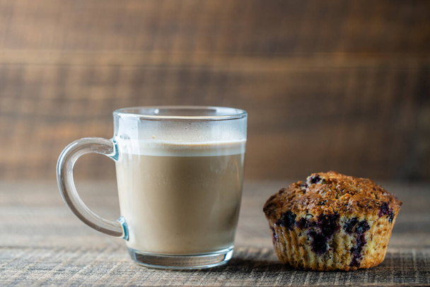 Delicious muffin with blueberries and cappuccino glass cup on a wooden table, close up. Fresh cupcake and coffee for breakfast - Фото, изображение