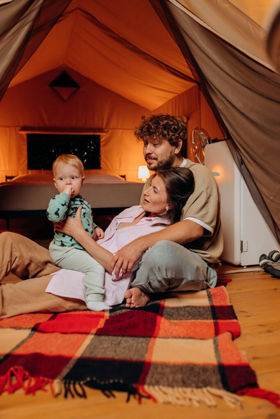 Glückliche Familie mit schönen Baby spielen und verbringen Zeit zusammen im Glamping am Sommerabend. Luxus-Zeltlager für Erholung und Erholung im Freien. Lifestyle-Konzept - Foto, Bild