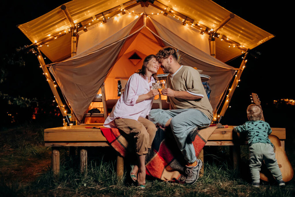 Couple heureux se détendre dans le glamping le soir d'été et boire du vin près du feu de joie confortable. Tente de camping de luxe pour les loisirs de plein air et les loisirs. Concept de style de vie - Photo, image