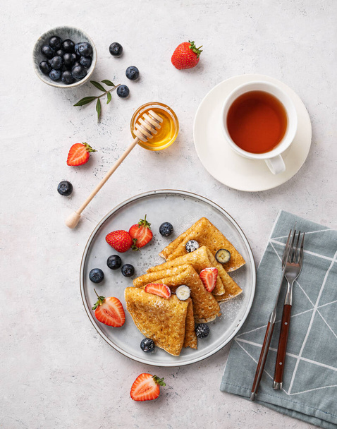 Homemade thin pancakes or crepes with fresh blueberries and strawberries, honey and cup of tea on a light background. Top view. - Foto, Imagem