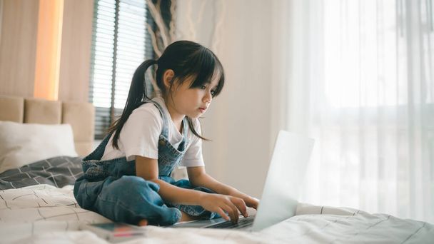 Asyalı kız çocuk yatakta oturuyor ve internette internetten öğreniyor. Evde yalnızlık çekerken videoyla ilgili sanal sınıf dersi. Uzaktan kumandalı video eğitimi. Çocuklar için modern okul eğitimi - Fotoğraf, Görsel