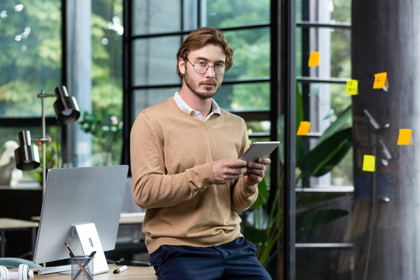 Портрет молодого студента-чоловіка, який стоїть в офісі, співпрацює за столом з комп'ютером, використовує планшет, навчається онлайн. Дивиться в камеру
. - Фото, зображення