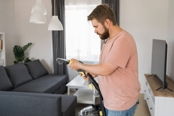Man holding modern washing vacuum cleaner and cleaning dirty sofa with professionally detergent. Professional springclean at home - Foto, Imagen