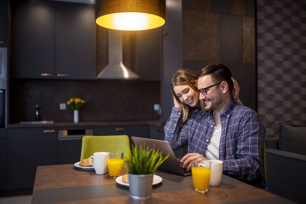 Picture of young couple looking at laptop on table while eating breakfast in kitchen at home. - Photo, Image