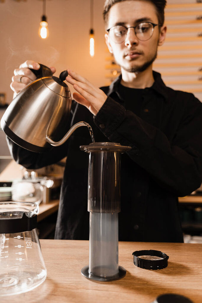 Barista is pouring hot water from drip kettle to aeropress. Process of brewing aeropress. Pouring hot water over roasted and ground coffee beans in aeropress - Foto, Imagen