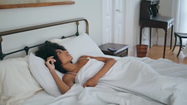 Соня просыпается в постели. Африканская американка проводит время в спальне, проверяя мобильный телефон. Кудрявая брюнетка просматривает новости смартфона, лежащие на уютном матрасе. Спокойная женщина просыпается в отеле - Кадры, видео