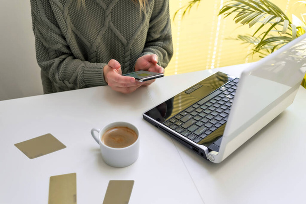Una mujer trabaja en una computadora portátil y mira la pantalla de su teléfono. El concepto de compras en línea, freelancing, aprendizaje, comunicación - Foto, imagen