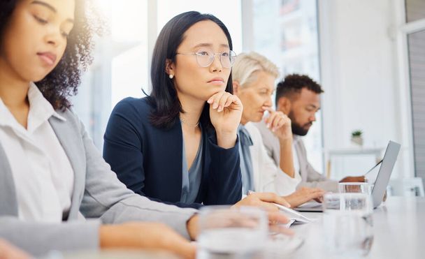 Serious young asian businesswoman wearing glasses and thinking with hand on her chin while listening to presentation meeting in an office boardroom with colleagues. Focused woman considering ideas an. - 写真・画像