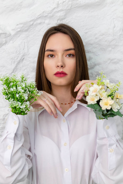 Portrait of a young beautiful brown-eyed girl in a white shirt, with flowers in her hands. A white wall is in the background. Beauty and Freshness concept. - Photo, Image