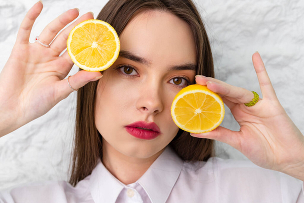 Porträt einer jungen schönen braunen Augen Mädchen in einem weißen Hemd hält und posiert mit runden Scheiben Zitrone. Im Hintergrund ist eine weiße Wand zu sehen. Schönheits- und Frischekonzept. - Foto, Bild