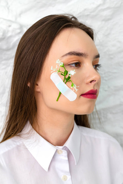 Close-up. Portrait of a young beautiful and cute brown-eyed girl with a flower attached to her cheek with a plaster. A white wall is in the background. Beauty and Freshness concept. - Photo, Image