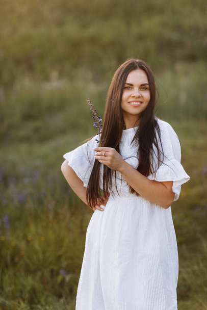 Genç, gülümseyen, beyaz elbiseli, mor lupin çiçekli güzel bir kadın baharda yeşil çayırda duruyor. Kadınlar Günü 'nde. Yaz zamanı. Romantik bir ruh hali. Doğa konsepti. - Fotoğraf, Görsel