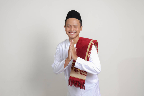 Πορτρέτο του ελκυστικού Ασιάτη μουσουλμάνος άνθρωπος σε λευκό πουκάμισο με skullcap δείχνει συγγνώμη και ευπρόσδεκτη χειρονομία χέρι. Μεμονωμένη εικόνα σε γκρι φόντο - Φωτογραφία, εικόνα