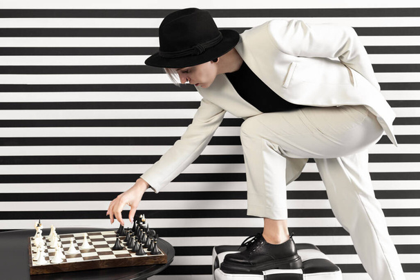 a beautiful woman in a strict white suit reaches out for one of the pieces on a chessboard with one leg on a chair. Side view. - Foto, Bild