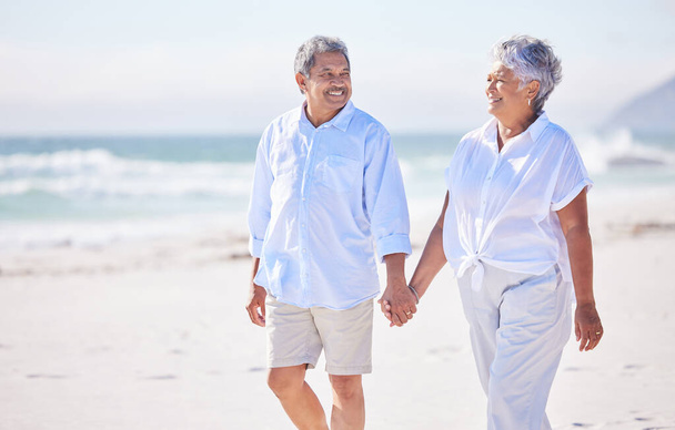 Happy mixed race senior couple holding hands while walking on the beach together. Mature couple traveling and enjoying retirement. - 写真・画像