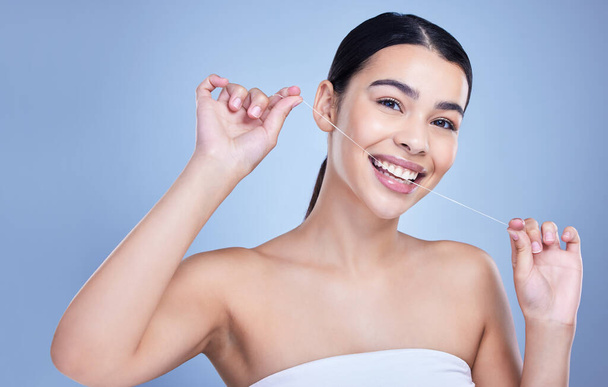 Studio portrait of a smiling mixed race young woman with glowing skin posing against blue copyspace background while flossing her teeth for fresh breath. Hispanic model using floss to prevent a cavit. - Fotoğraf, Görsel