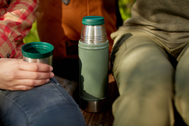 Female hiker drinking a mug of hot coffee from a thermal flask on the bench alongside her as she sits chatting to a friend - Photo, image
