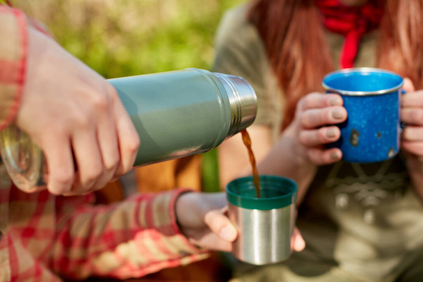Young woman serving herself hot coffee from a thermal flask as she relaxes outdoors with a girl friend while out hiking or camping - Photo, image