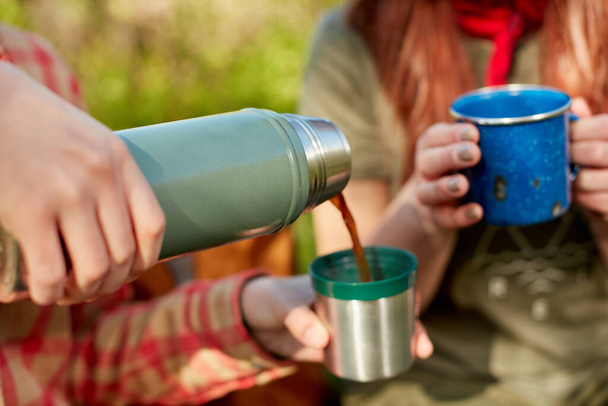 Two women enjoying hot coffee on a hike pouring it from a thermal flask into mugs in a close up view of their hands - Foto, Imagem