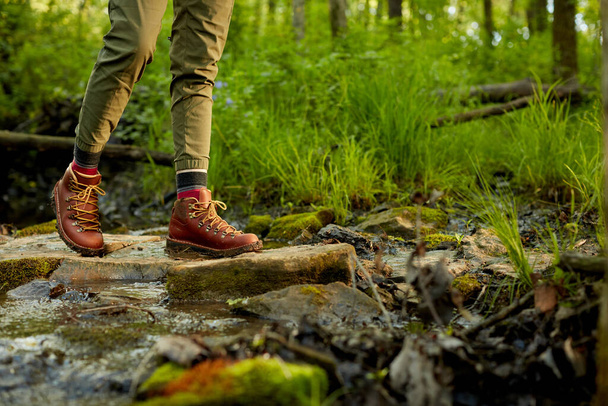 Woman hiking across a small rocky stream bordered by lush green grass in a close up view of her brown leather boots - 写真・画像
