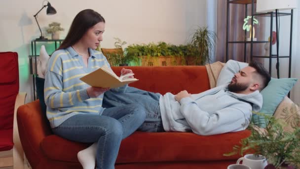 Woman reading story from book while displeased tired boyfriend man falling asleep and snoring. Young family couple at home. Husband wife together on sofa in living room. Hobby leisure activities - Footage, Video