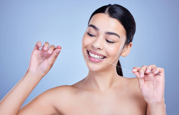 Eine fröhlich lächelnde junge Frau mit glühender Haut posiert vor blauem Hintergrund, während sie die Zähne zusammenbeißt, um frischen Atem zu bekommen. Hispanisches Modell mit Zahnseide, um einen Hohlraum zu verhindern. - Foto, Bild