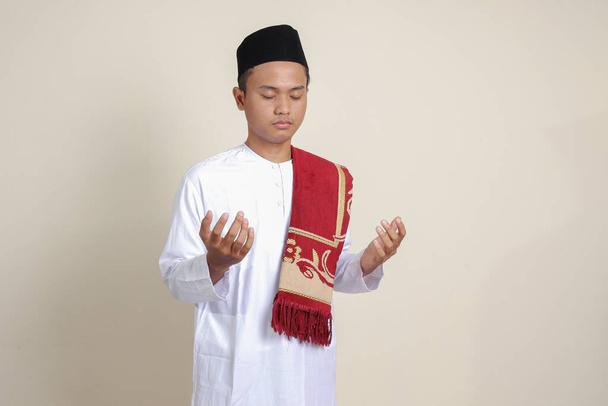 Beyaz gömlekli yakışıklı Asyalı Müslüman adamın portresi. Kafatası şapkalı. Elleri havada dua ediyor. Gri arkaplanda izole edilmiş resim - Fotoğraf, Görsel