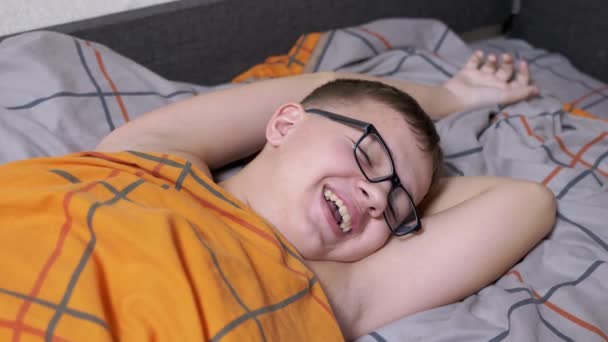 ベッドの上に寝そべっている眼鏡の中の眠そうな笑顔の男の子,口を開けて,八幡,ストレッチ.柔らかい居心地の良いベッドで、オレンジの毛布で覆われ、目を閉じて休んで魅力的な10代の疲れ。寝る時間だ。リラクゼーション. - 映像、動画