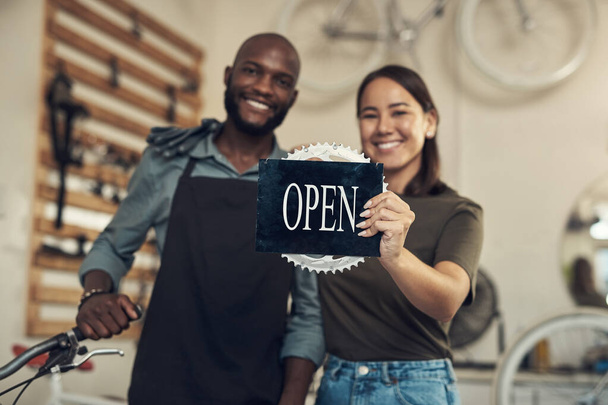 Siéntase libre de entrar. dos jóvenes propietarios de negocios de pie juntos en su tienda de bicicletas y sosteniendo un cartel abierto - Foto, imagen