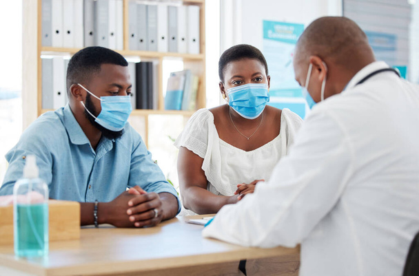 Young couple wearing covid masks during medical consult.Specialist talking to married couple about ivf treatment. African american couple in checkup for fertility. Expert doctor talking to patients. - Фото, изображение