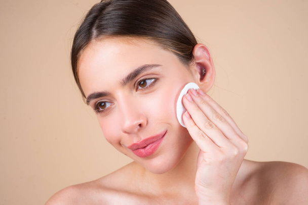 Close up beautiful young woman with cotton pad removing makeup. Skin care. Studio portrait of girl cleaning her face with cotton pad, isolated beige background. Face toner and cotton pad - Foto, afbeelding