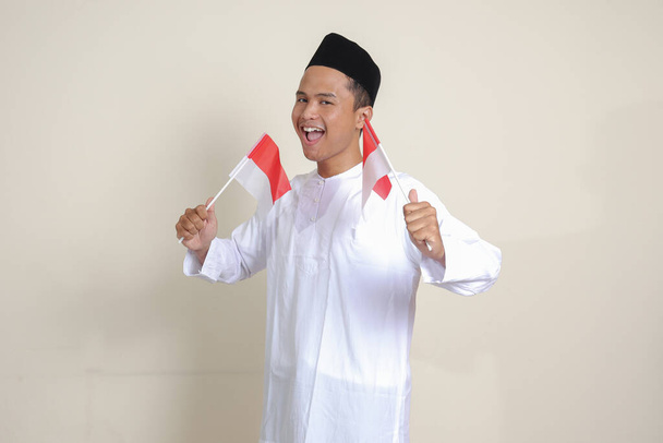 Portrait of attractive Asian muslim man in white shirt with skullcap holding indonesia flag while raising his fist, celebrating success. Isolated image on gray background - Photo, Image