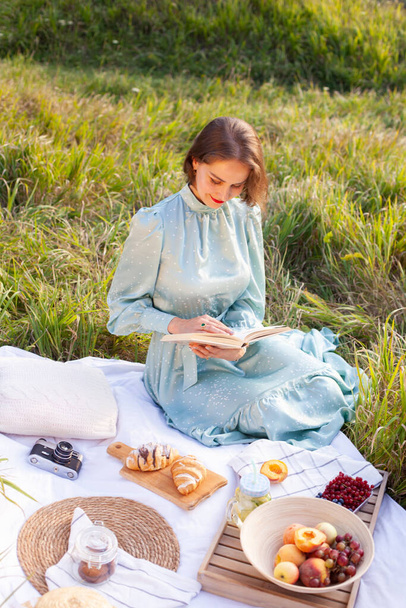 A woman in a long summer dress with short hair sitting on a white blanket with fruits and pastries and reading the book. Concept of having picnic in a city park during summer holidays or weekends.  - Foto, immagini