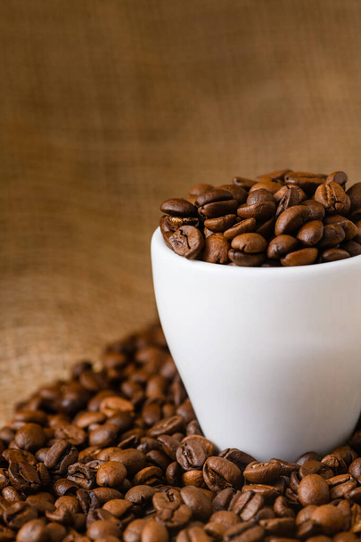 Taza completa de granos de café tostados en mesa cubierta por saco, colores cálidos naturales, bebida de la mañana - Foto, Imagen